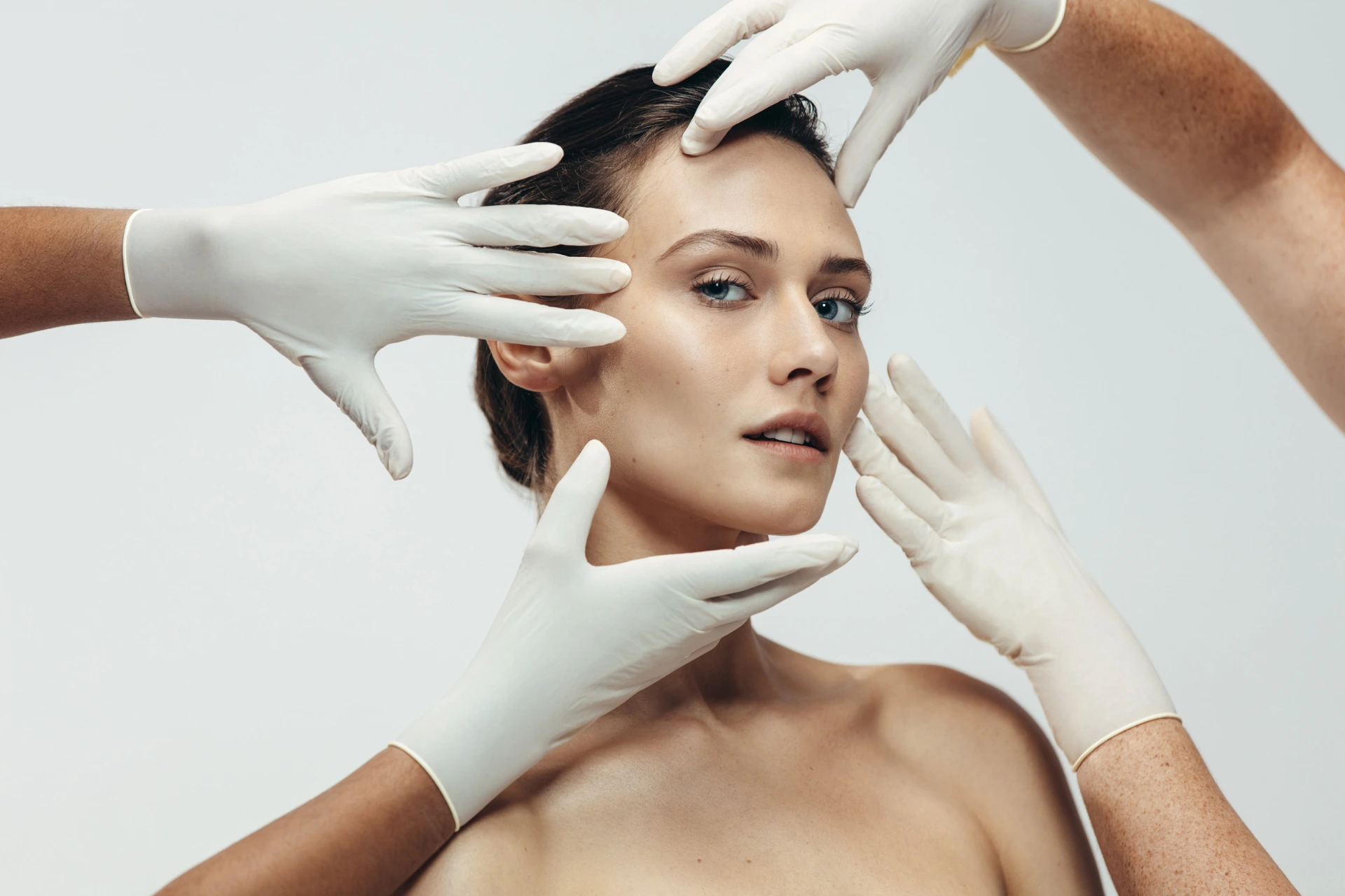Mujer con piel radiante y manos con guantes evaluando su rostro, representando tratamientos estéticos personalizados.